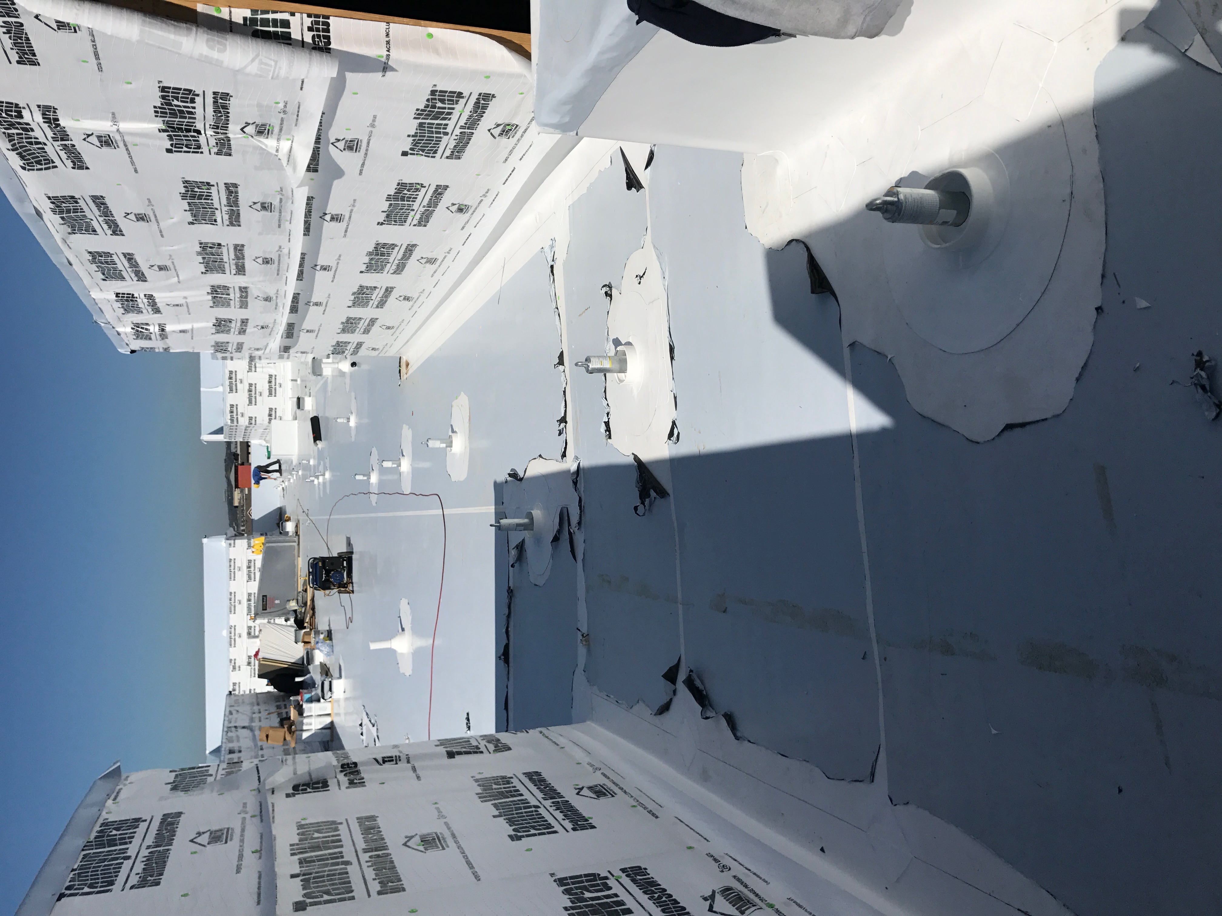 White TPO commercial roof installation in progress showing fastener plates, insulation boards and TamlynWrap wall underlayment with a crew member in the distance