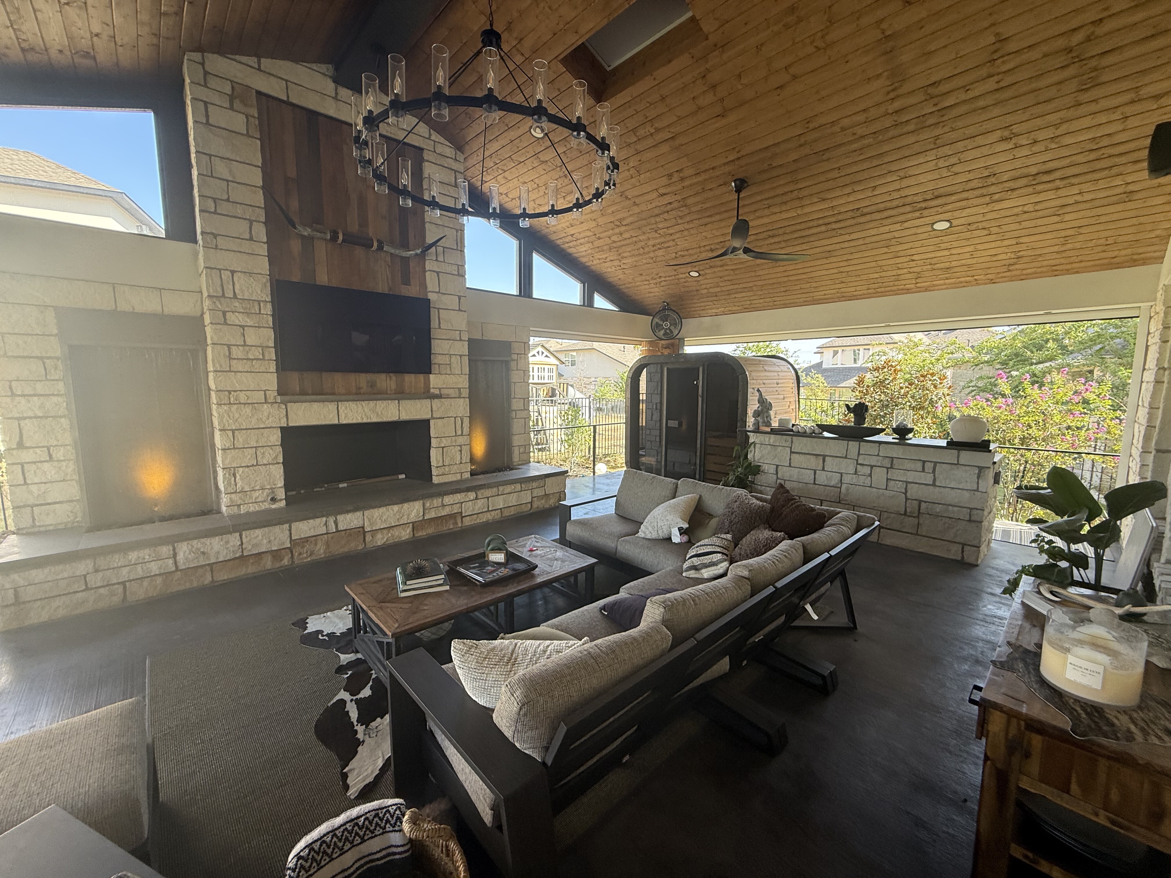 Covered outdoor living room with stone fireplace, wood ceiling, chandelier, sofa seating, sauna and outdoor bar designed as one unified space
