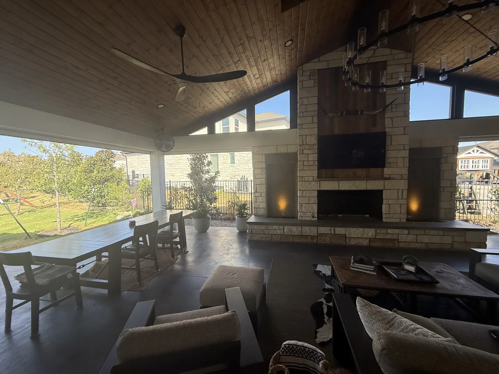 Wide covered living and dining space with lounge seating facing a stone fireplace and mounted television