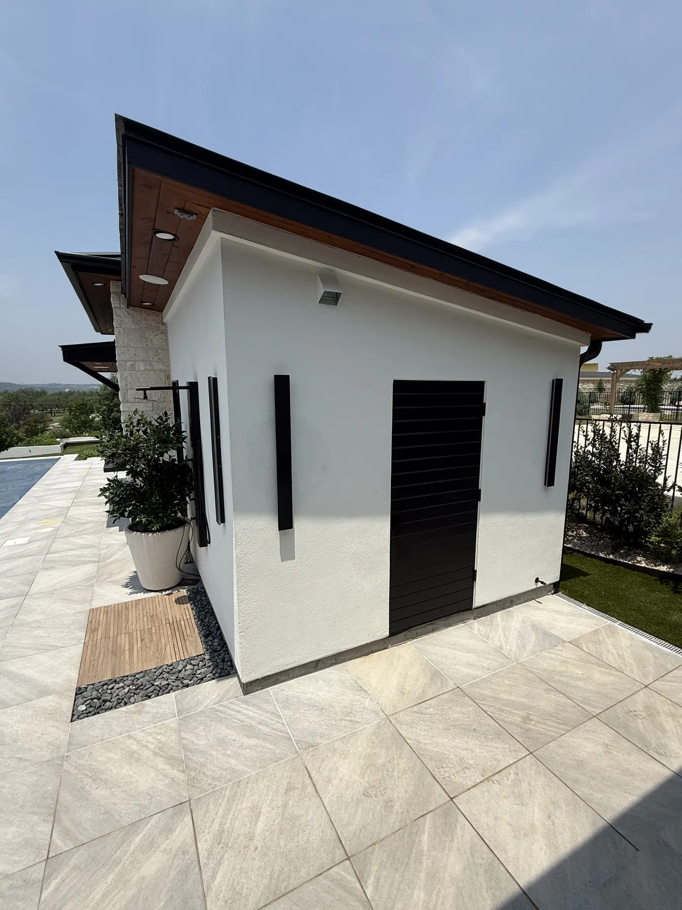Detached white structure with black louvered doors and a wood-clad soffit standing beside a pool on a tiled terrace