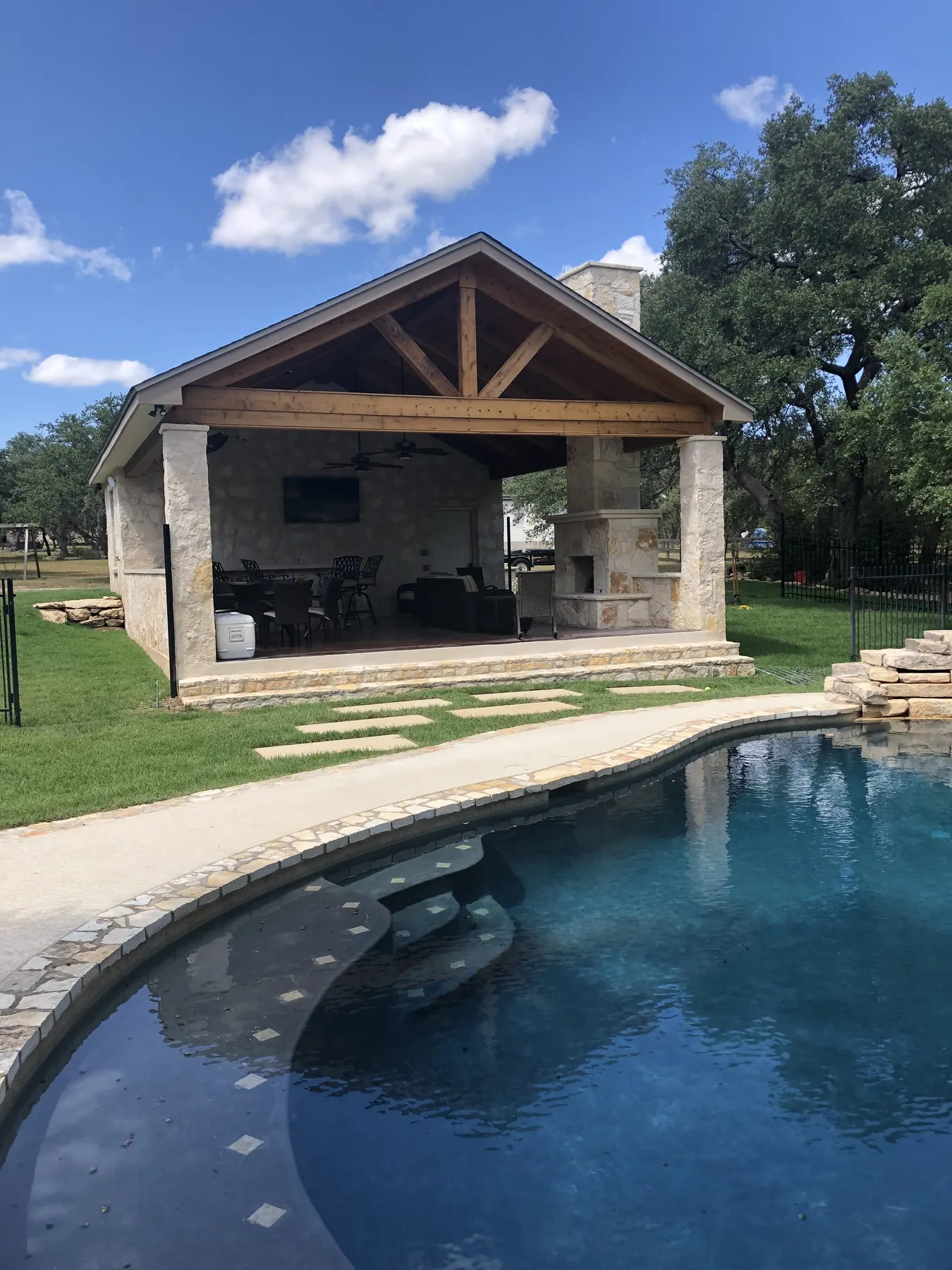 Freestanding stone pavilion with a timber gable roof beside a swimming pool and mature oaks