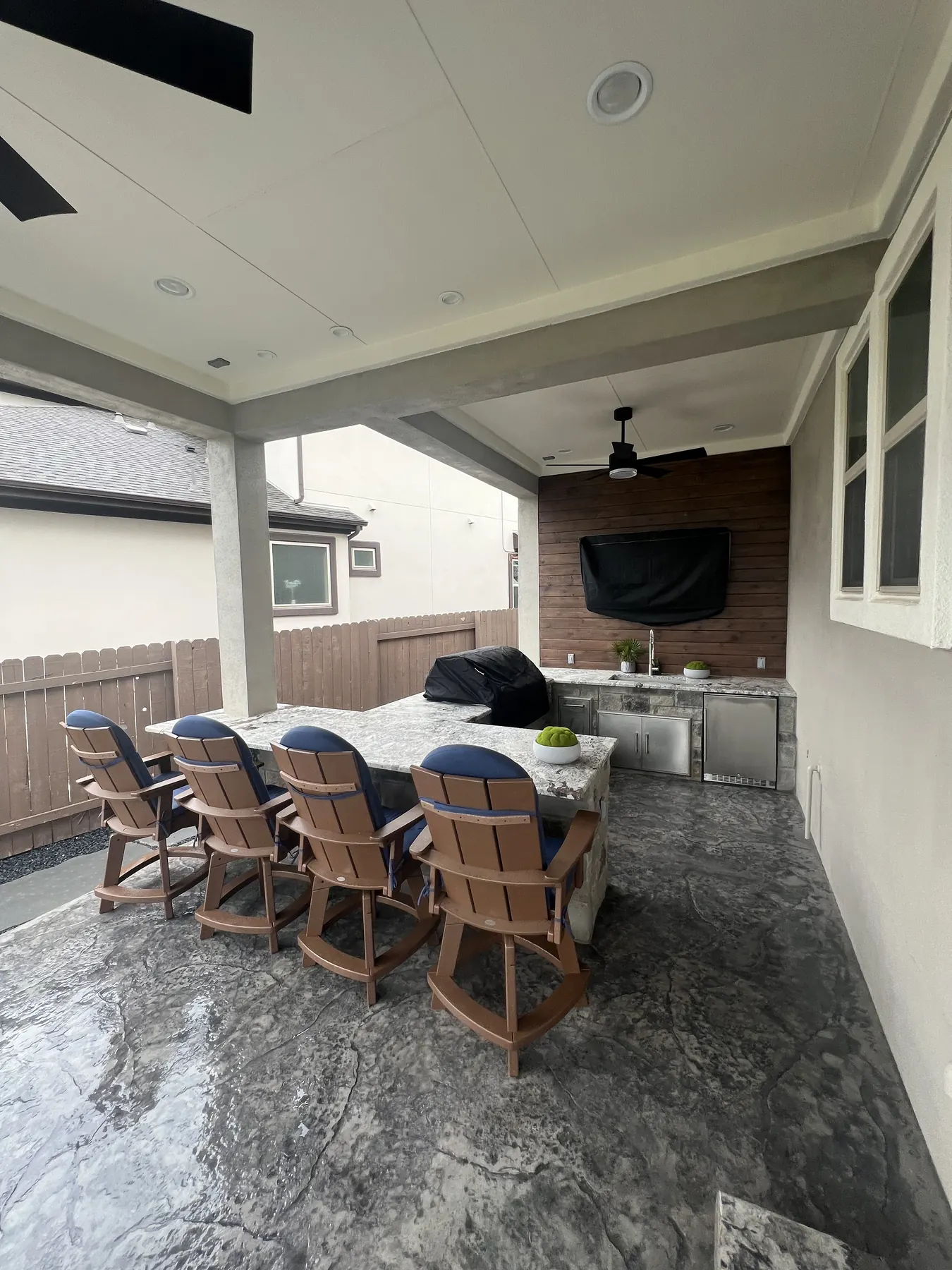 Covered patio with a stone bar counter, barstools, mounted television and outdoor kitchen along the rear of the home