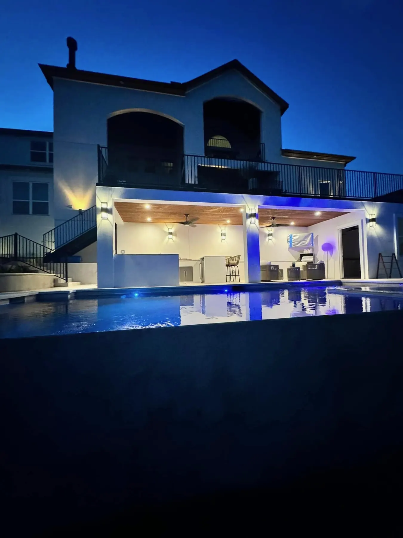 Pool and covered outdoor kitchen lit in the evening beneath a two-story home with a balcony