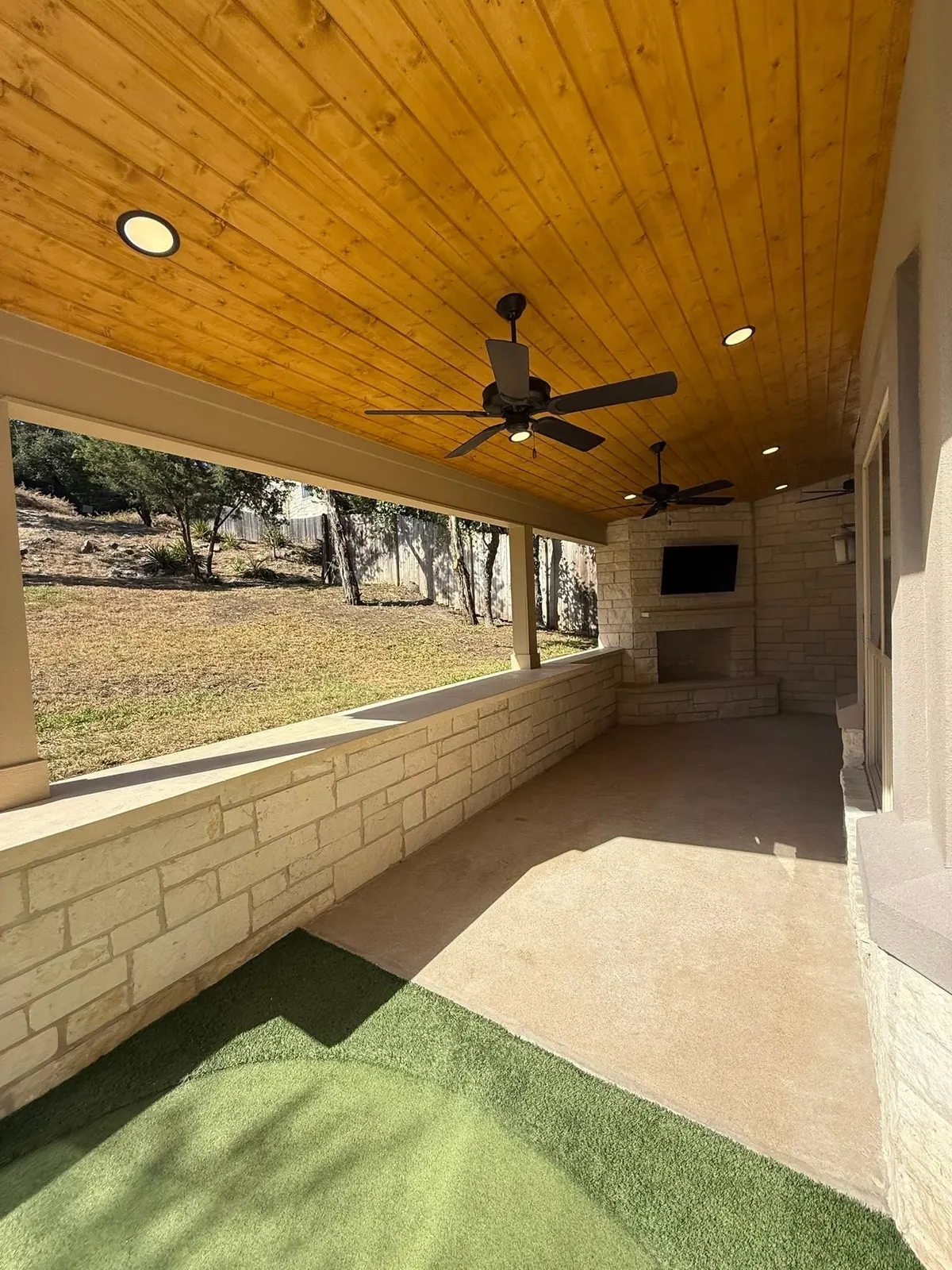 Covered patio with a warm wood ceiling and stone wall opening onto a turf lawn