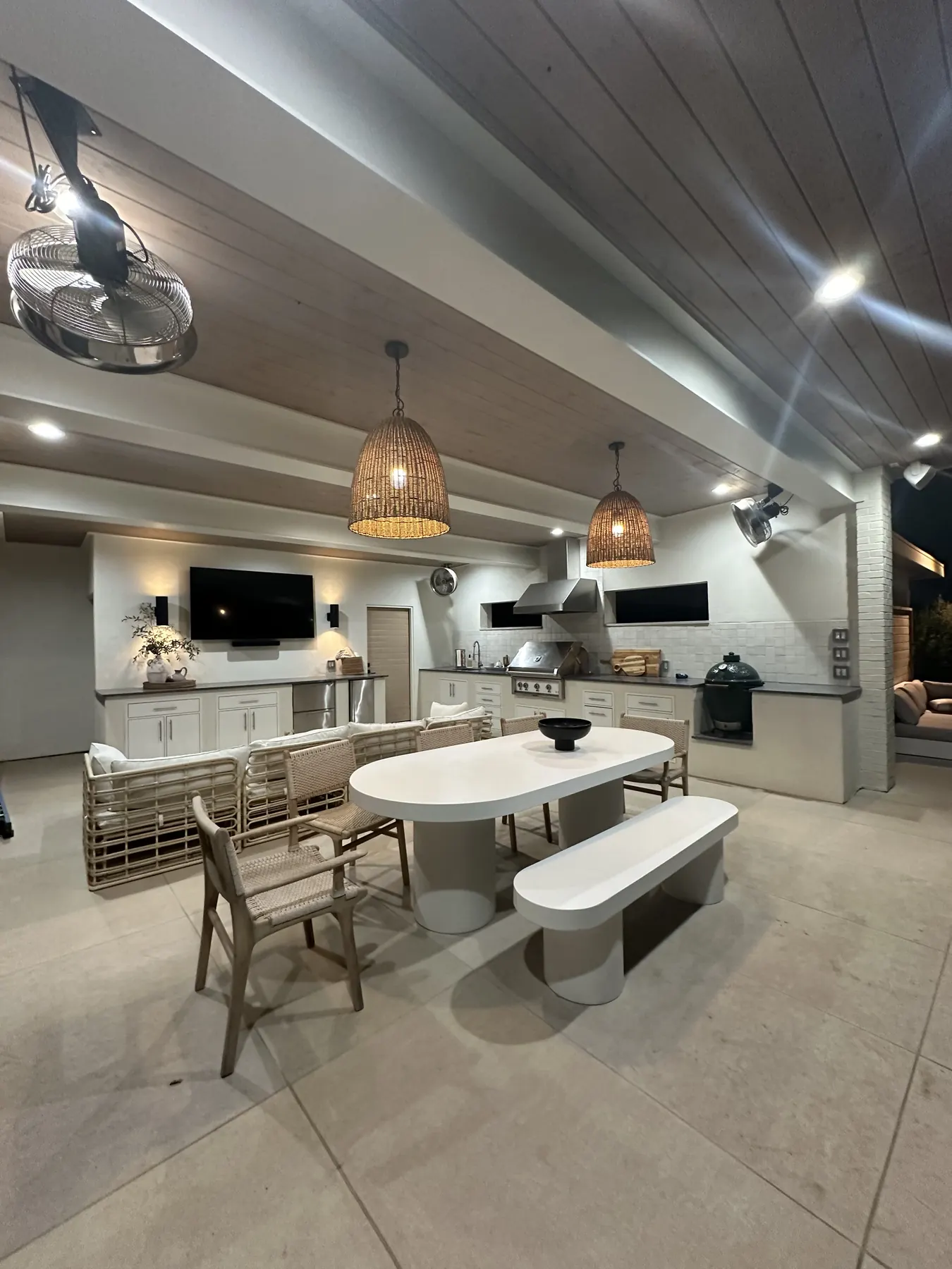 Covered outdoor kitchen and dining space with a stained wood ceiling, woven pendant lights and a long stone dining table